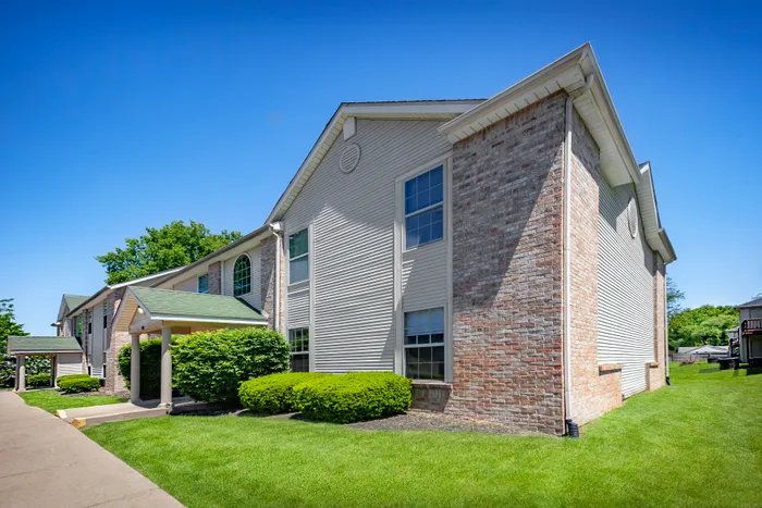 Charming two-story apartment building with a classic brick facade, surrounded by lush greenery and a bright blue sky.