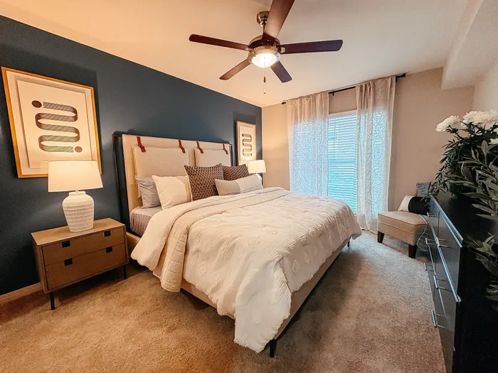 River Crossing elegant bedroom with a king-sized bed, plush pillows, and cozy lighting. Features stylish artwork, a ceiling fan, and large windows with soft drapes for natural light.