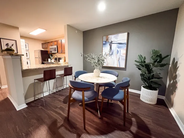 River Crossing modern dining area with elegant seating for four, featuring stylish artwork, lush greenery, and a glimpse of the contemporary kitchen with sleek bar stools.
