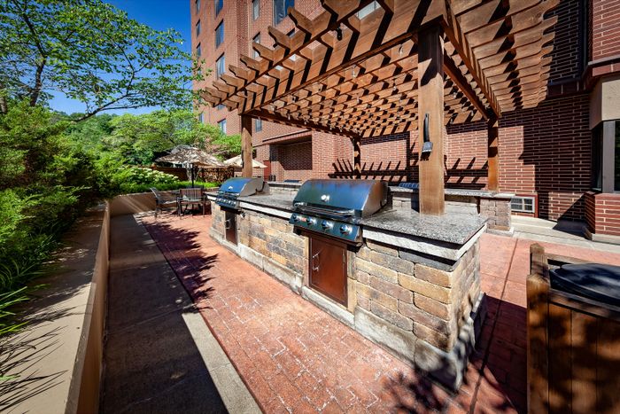 Chic outdoor barbecue area with twin grills under a stylish wooden pergola, surrounded by lush greenery and brick exterior, perfect for entertaining.