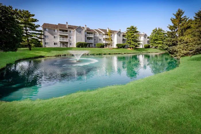 Scenic apartment community with serene pond and fountain, surrounded by lush greenery and charming residences.