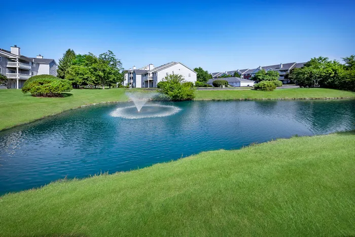 Serene apartment community with scenic pond view featuring a tranquil fountain, lush green lawns, and modern white buildings against a clear blue sky.