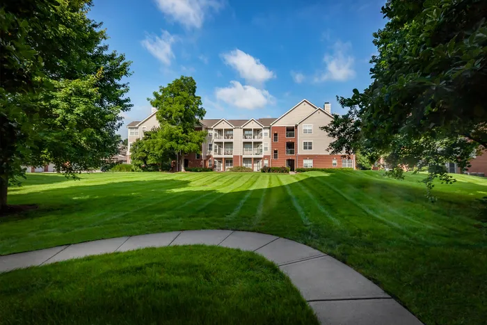 Charming apartment complex with spacious balconies, surrounded by lush green lawns and mature trees, under a bright blue sky.