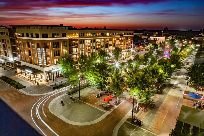 Railyard at Midtown Apartments - Vibrant evening view of a lively urban neighborhood, showcasing modern architecture, tree-lined streets, and dynamic street lighting under a colorful sunset sky.