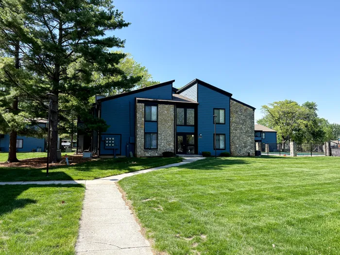 Charming suburban apartment building with unique stone and dark blue siding, surrounded by lush green lawns and tall trees under a clear blue sky.