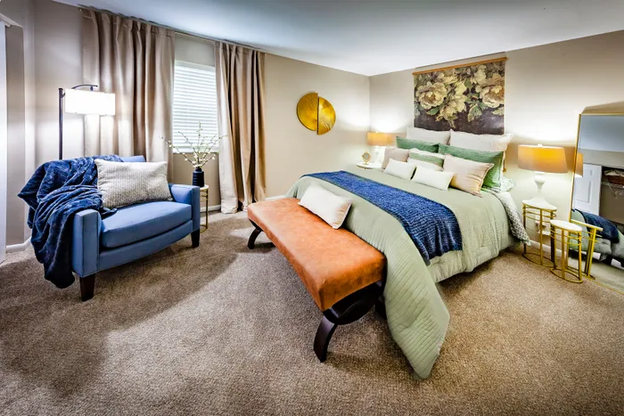 Elegant bedroom with a plush green and blue bed setup, cozy armchair, and vibrant wall art, exuding warmth and sophistication.
