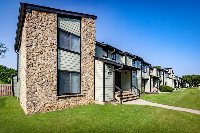 Pebble Point Apartments - Charming residential building with rustic stone accents and modern pastel siding, set in a lush green landscape under a clear blue sky.