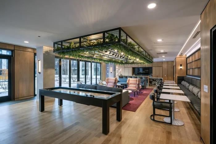The Elex Apartments - Modern communal lounge area featuring chic seating, a green ceiling installation, and a game table with bright natural lighting.