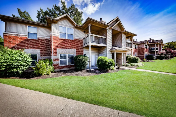 Charming apartment complex with brick facade, lush green lawns, and spacious balconies under a bright blue sky.