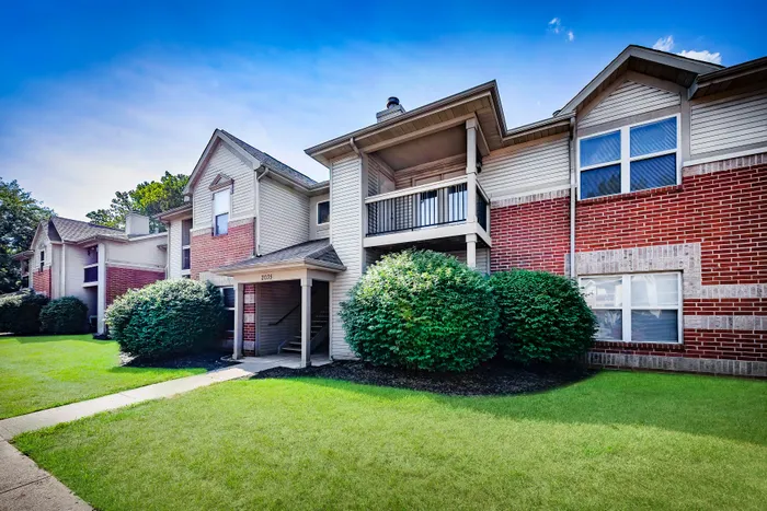 Charming brick apartment complex with lush greenery and spacious balconies, set under a picturesque blue sky.