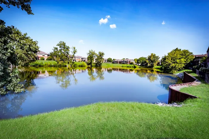 Scenic view of peaceful apartment complex nestled by a tranquil lake surrounded by lush greenery under a bright blue sky.