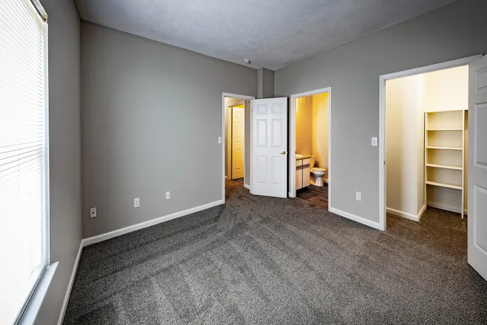 Spacious bedroom with plush carpet, showcasing an open closet and an en-suite bathroom, filled with natural light through large windows.