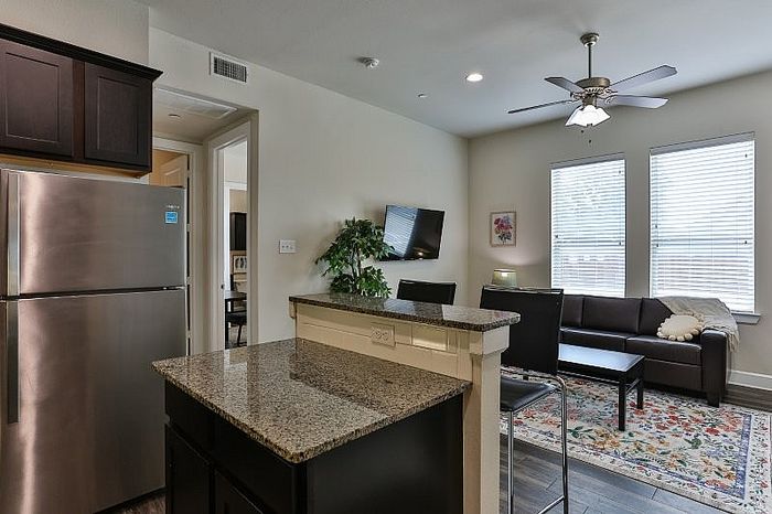 Modern apartment kitchen with granite countertops, stainless steel appliances, and an open-concept living area featuring a ceiling fan and cozy seating.