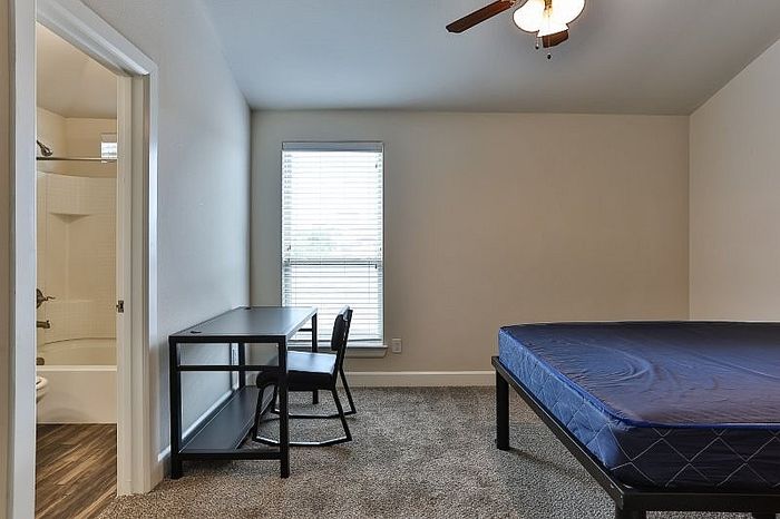Modern bedroom with cozy carpeted flooring, featuring a comfortable bed and a sleek desk by a sunlit window, adjacent to an en-suite bathroom.