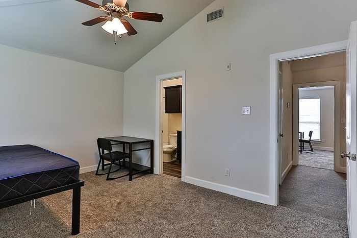 Bright bedroom with plush carpet, elegant ceiling fan, and an en-suite bathroom, creating a cozy and functional living space.