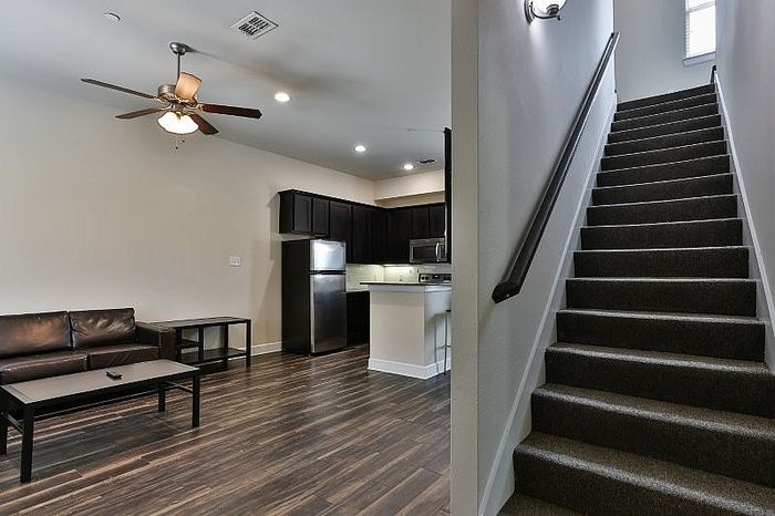Elegant living area with modern kitchen, featuring dark wood flooring and sleek stainless steel appliances, beside a stylish carpeted staircase leading to upper levels.