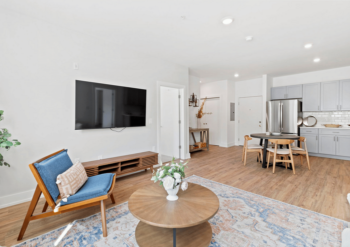 Bright open living space featuring a modern lounge chair, wall-mounted TV, round dining table, and sleek kitchen in a cozy apartment.