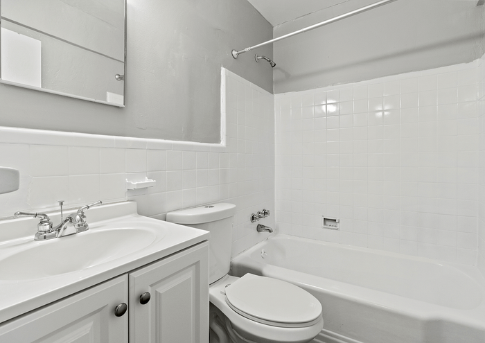 Brightly lit bathroom with white tiles featuring a bathtub, vanity with sink, and a large mirror.