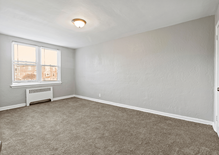 Sunlit empty room with neutral walls and carpet, featuring large windows and a ceiling light.
