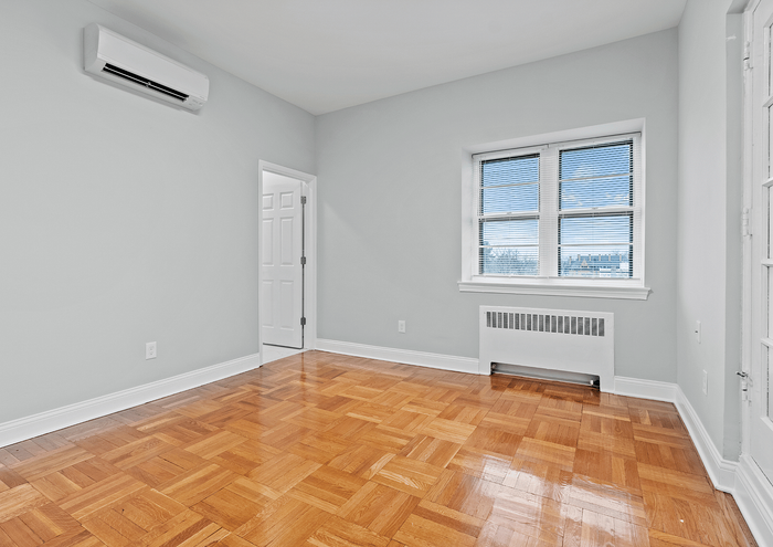 Spacious, sunlit room with polished wooden floors and a large window offering city views.