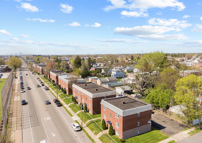 Aerial view of a suburban street with red brick apartment buildings, lush green trees, and a distant city skyline.