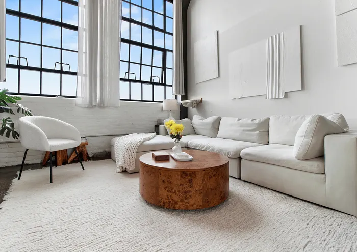 Beautiful loft-style living room with large industrial windows, comfortable white sectional sofa, and round wooden coffee table.