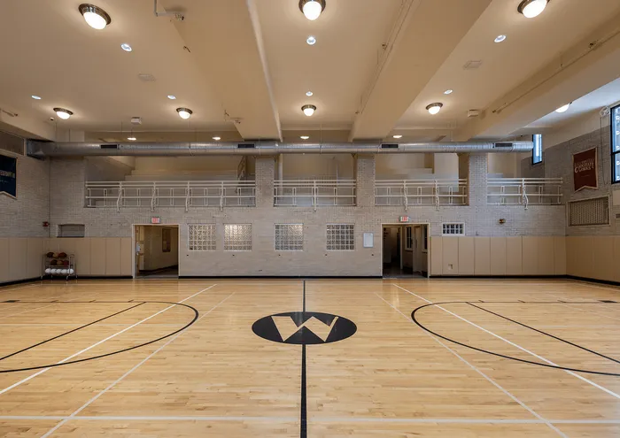 Spacious indoor gymnasium with polished hardwood floors, bright lighting, and upper balcony area.