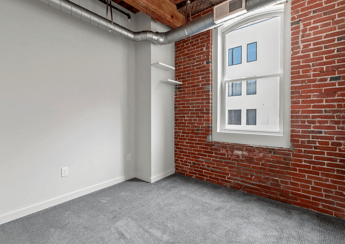 Bright room with exposed brick walls and industrial ductwork, featuring a large window and modern gray carpet.