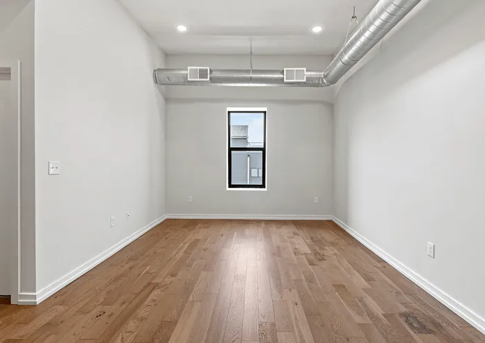 Bright, empty room with hardwood floors, modern ductwork, and a large window.