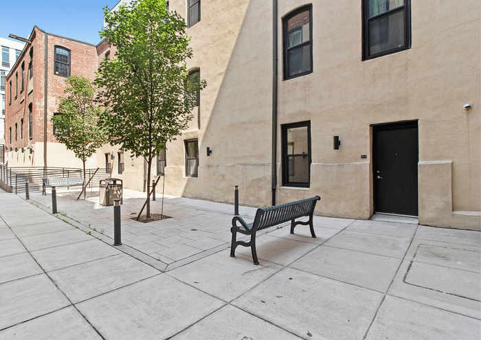 Welcoming apartment entryway with brick facade, trees, and a bench in a sunlit courtyard.