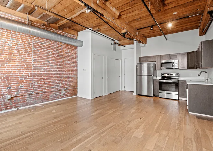 Modern loft apartment featuring exposed brick wall, wooden beams, open kitchen with stainless steel appliances, and hardwood floors.