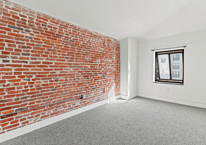 Stylish apartment room featuring an exposed brick wall, large window, and modern gray carpeting.