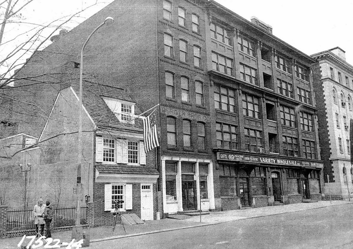 Vintage city street view featuring a small colonial house next to a large brick warehouse.