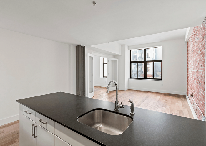 Bright, open-concept apartment kitchen and living area with large windows and exposed brick wall.