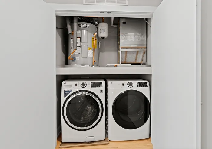 Modern laundry closet with front-loading washer and dryer beneath a water heater and air duct system.