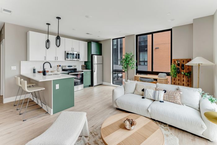 Modern apartment interior with an open-plan kitchen and living room featuring sleek white and green cabinetry, elegant lighting, and a cozy sofa next to large windows.