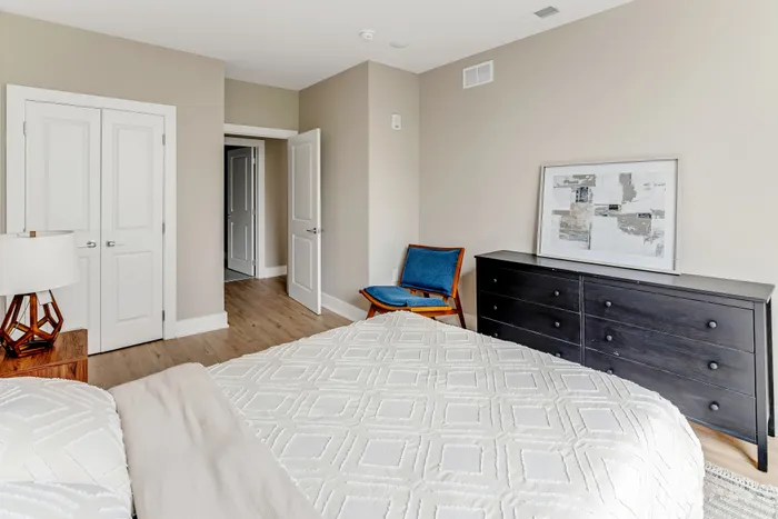 Elegant bedroom with modern decor featuring a cozy bed, stylish chair, and chic dresser, accented by contemporary art and warm, natural light.