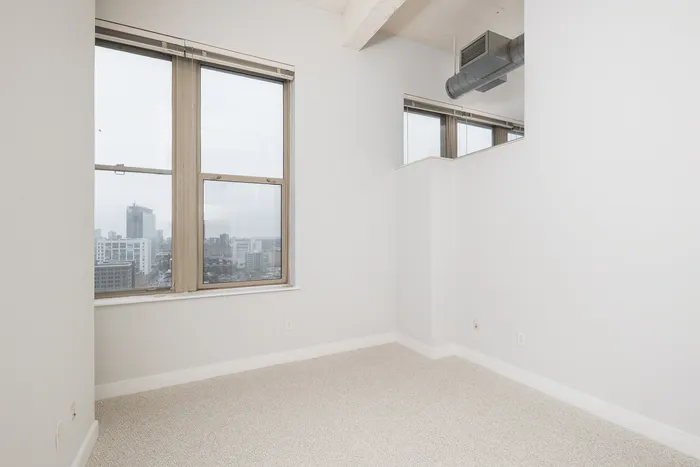Bright urban apartment with large windows showcasing a stunning city skyline view, freshly painted walls, and cozy carpeted floors.