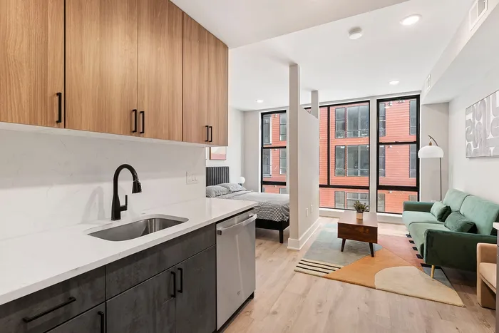 Modern studio apartment featuring a sleek kitchen with wood cabinetry, open living space with stylish furnishings, and abundant natural light from floor-to-ceiling windows.