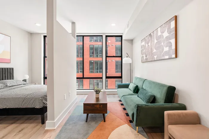 Modern studio apartment with stylish decor, featuring a cozy bed area separated by a partition, a chic green sofa, and large windows offering abundant natural light.