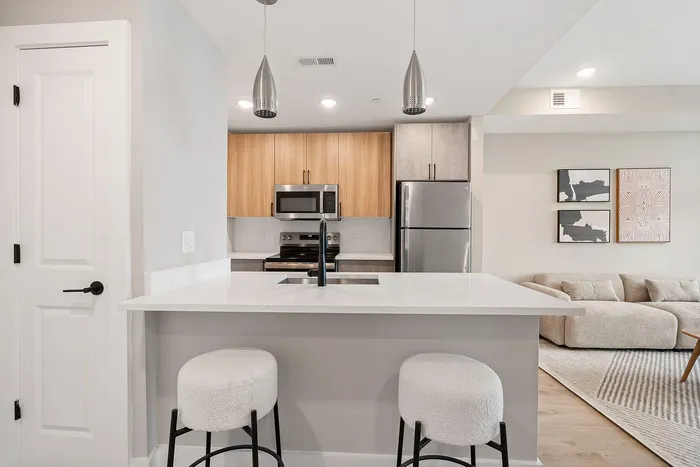 Modern kitchen with sleek stainless steel appliances, elegant pendant lighting, and a cozy breakfast bar with plush stools, adjacent to a stylish living area featuring a contemporary beige sofa.