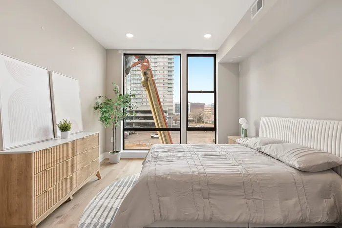 Bright and modern bedroom with large windows, elegant furnishings, and city views, featuring a cozy bed and stylish dresser.