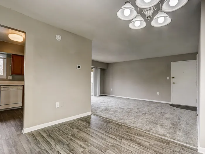 Stylish open-plan living area featuring sleek hardwood flooring and elegant chandelier, seamlessly connecting to a cozy carpeted space, with a glimpse of a functional kitchen.