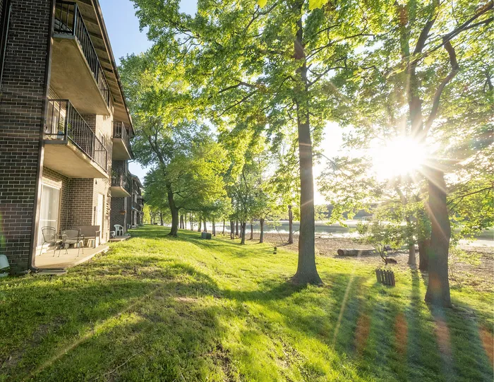 Charming apartment balcony view with lush green lawn, tall trees, and radiant sunlight peeking through, offering a serene lakeside atmosphere.
