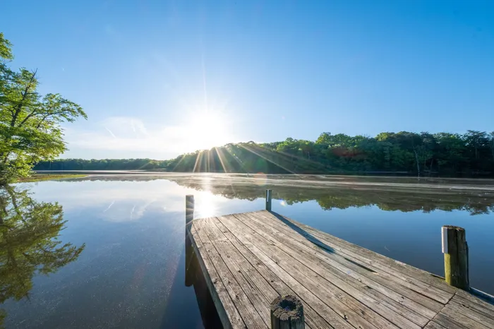 Scenic lakeside view with a wooden pier, calm waters, and a radiant sunrise over lush green trees.