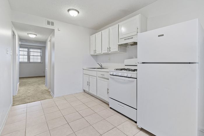 Bright and airy apartment kitchen with sleek white cabinetry, modern appliances, and an inviting hallway leading to natural light-filled rooms.