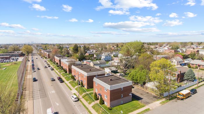 Charming suburban neighborhood with brick apartment buildings, tree-lined streets, and a distant city skyline under a bright blue sky.