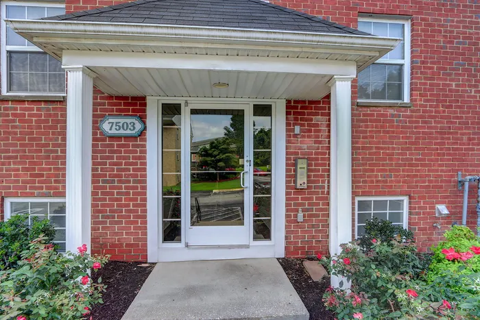 Charming brick apartment entrance with elegant awning, surrounded by lush greenery and vibrant flowers.