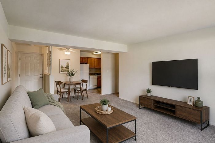 Bright and inviting living room with modern furniture, large wall-mounted TV, and an adjacent dining area leading to a cozy kitchen.