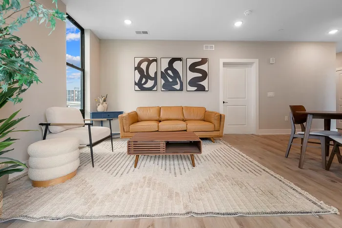 Bright and inviting living room with sleek decor, featuring a tan leather sofa, modern art, and expansive windows offering city views.